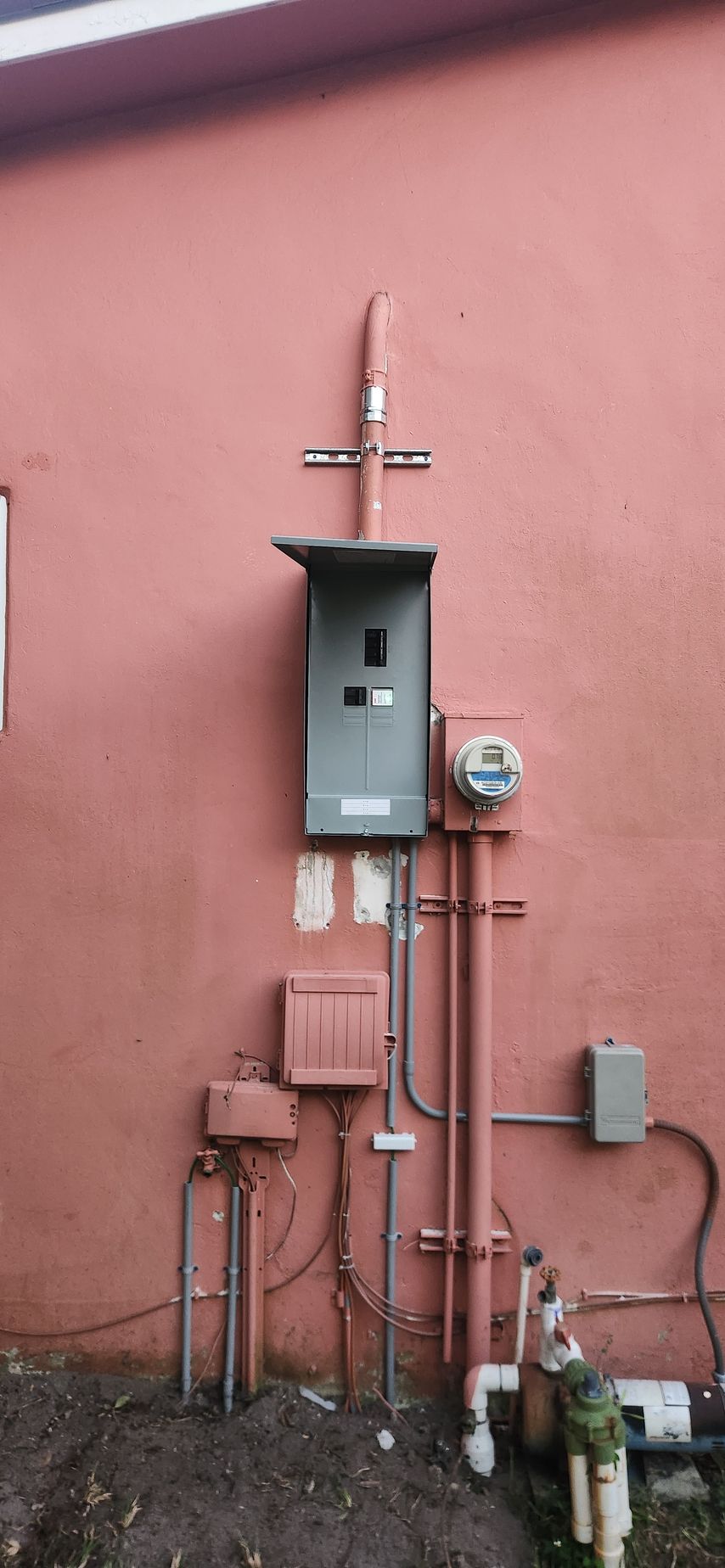 Outdoor utility meter and electrical boxes mounted on a pink wall with pipes and cables.