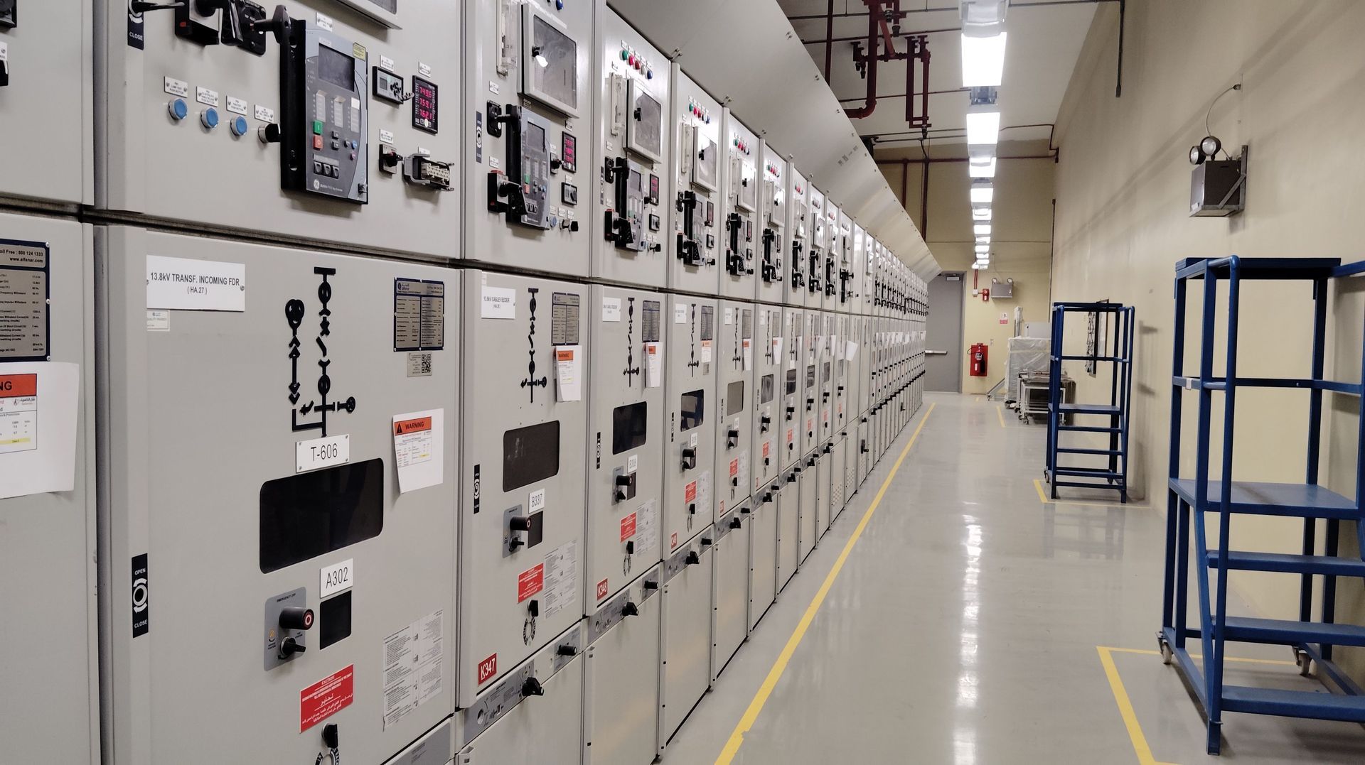 Long row of white electrical control panels in an industrial hallway with blue carts on the right.