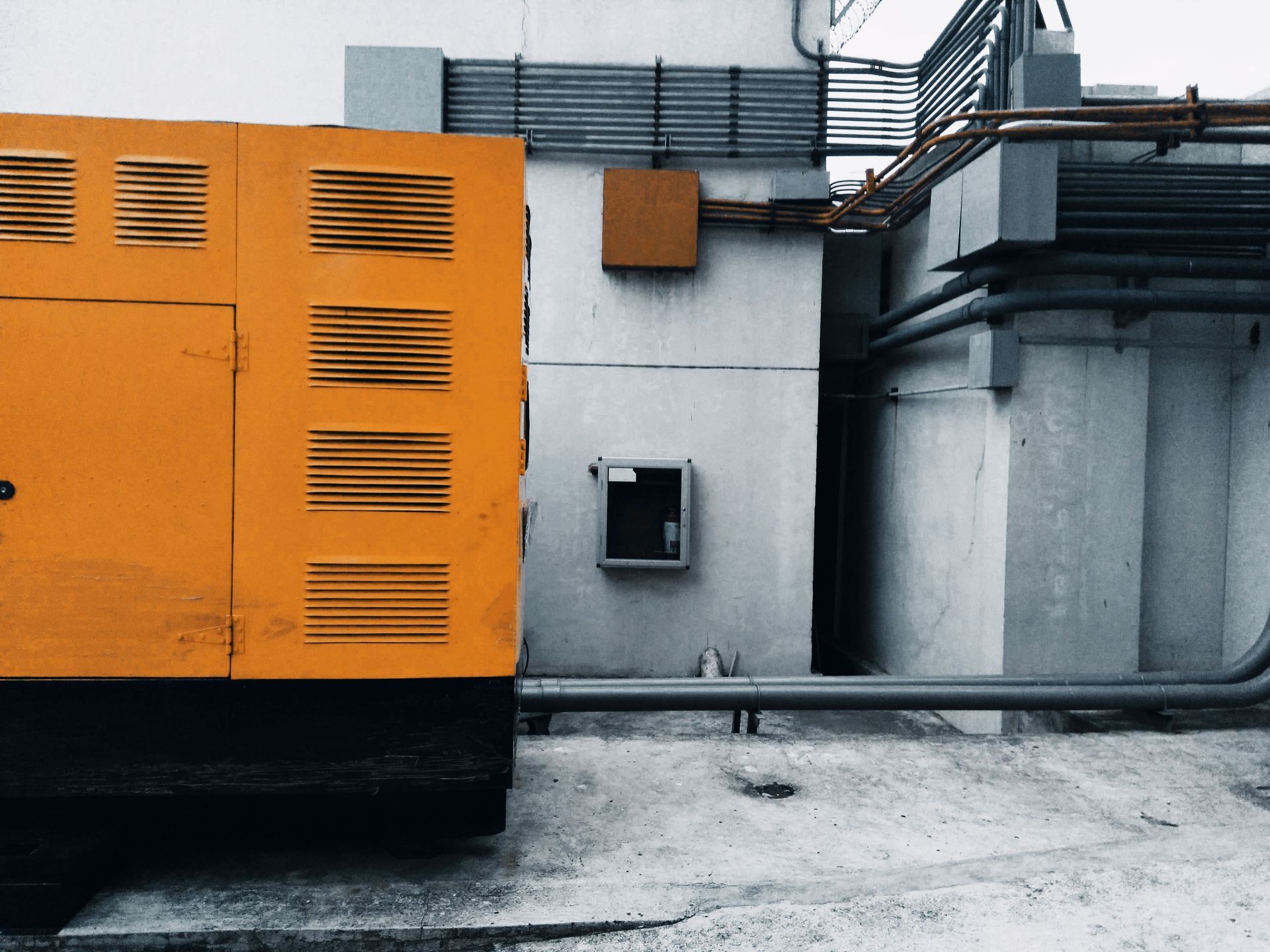 Orange industrial generator beside a gray building with pipes and vents.