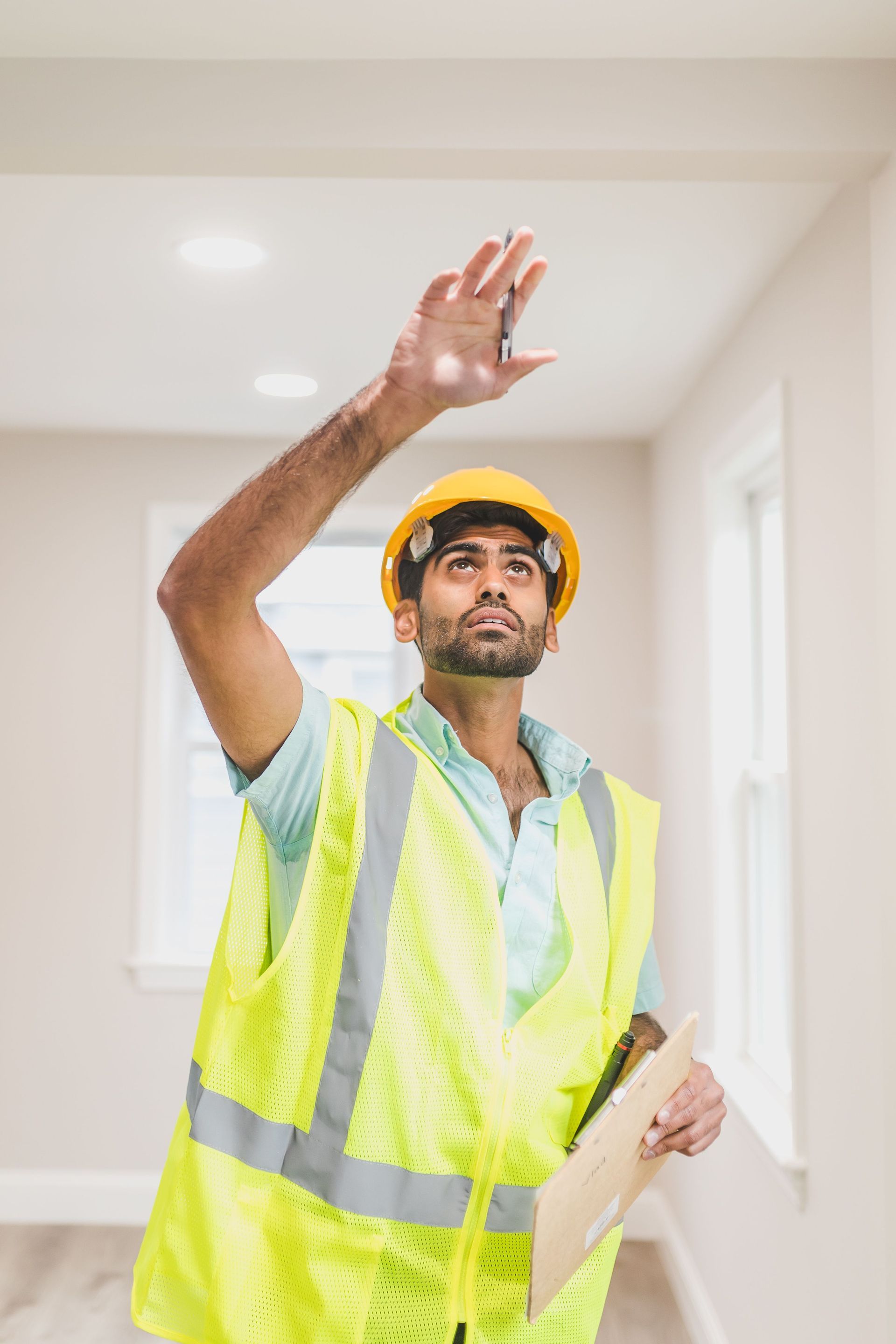 Construction worker in yellow hard hat and safety vest waving indoors while holding a clipboard
