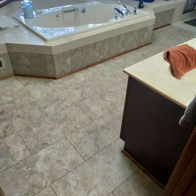 A bathroom features a large, off-white jetted bathtub with marble-look tile surround and matching square floor tiles.