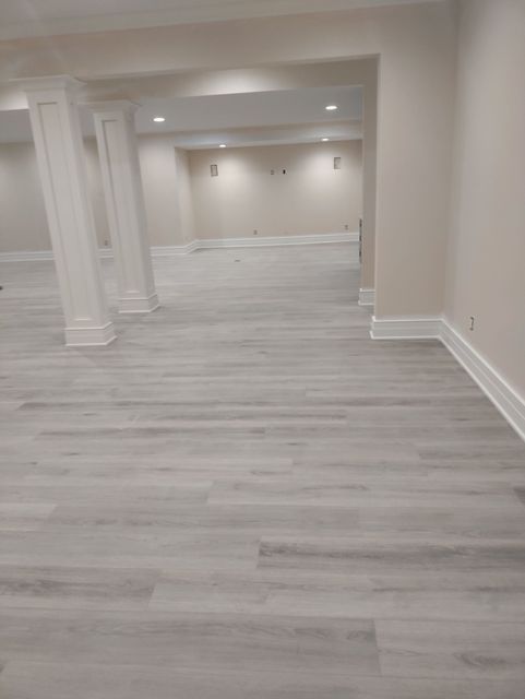 A modern basement interior with light wood floors, beige walls, white trim, and two square support columns.