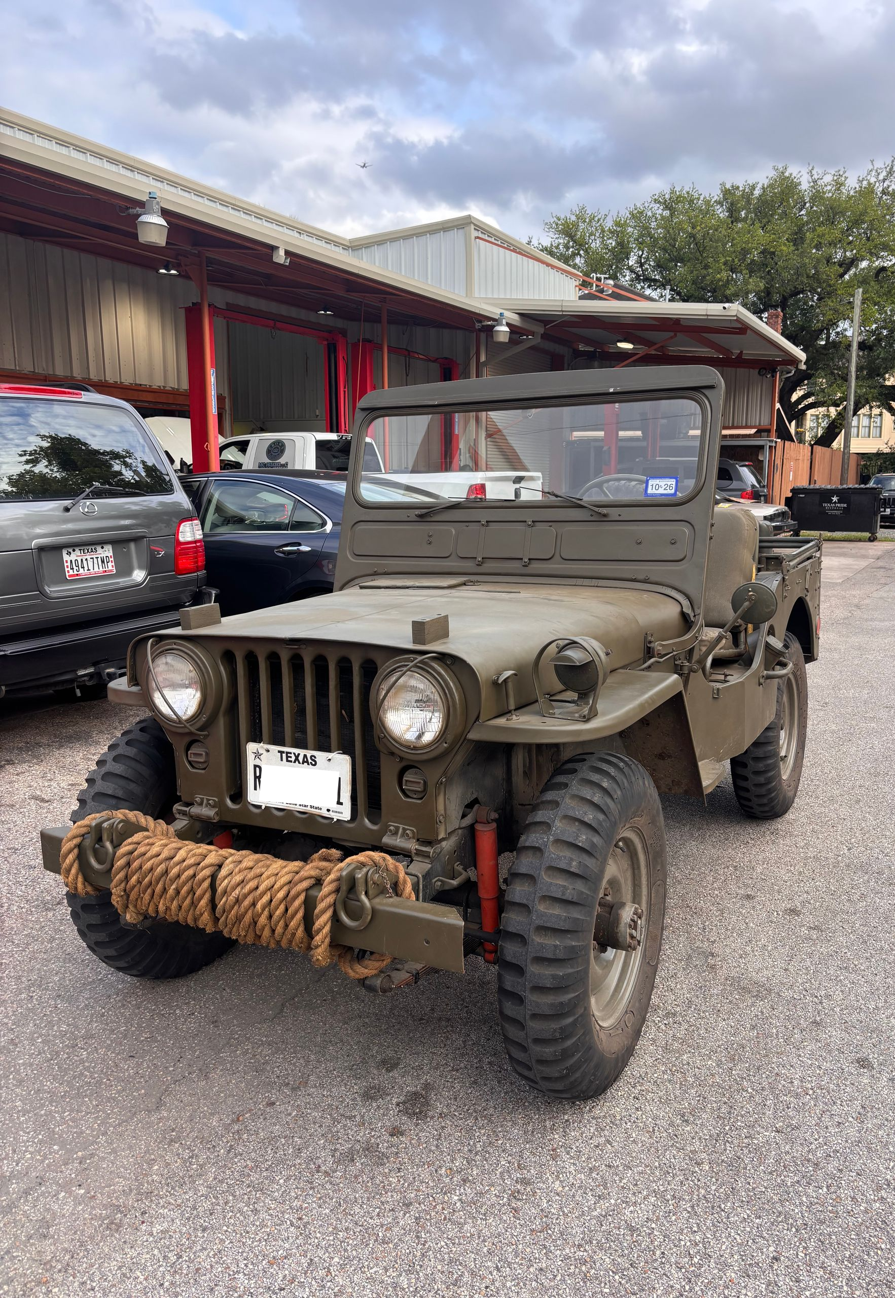 Vintage olive drab military jeep parked on a gravel lot beside other vehicles and a building.