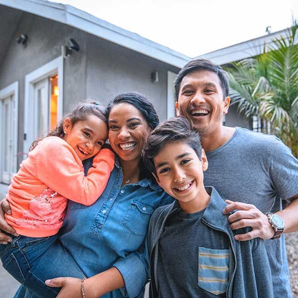 Smiling family of four posing outside a house, with one child leaning on a woman’s shoulder.