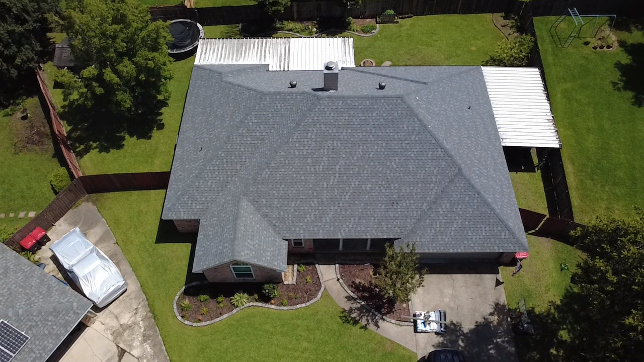 Aerial view of a house with a gray roof, green lawn, driveway, and surrounding trees.