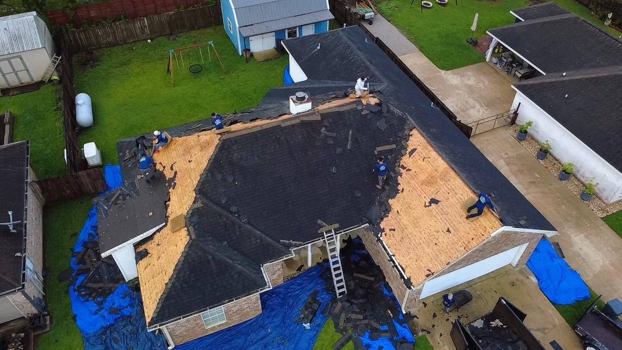 Damaged roof under repair, with exposed wood and blue tarps around a house in a neighborhood