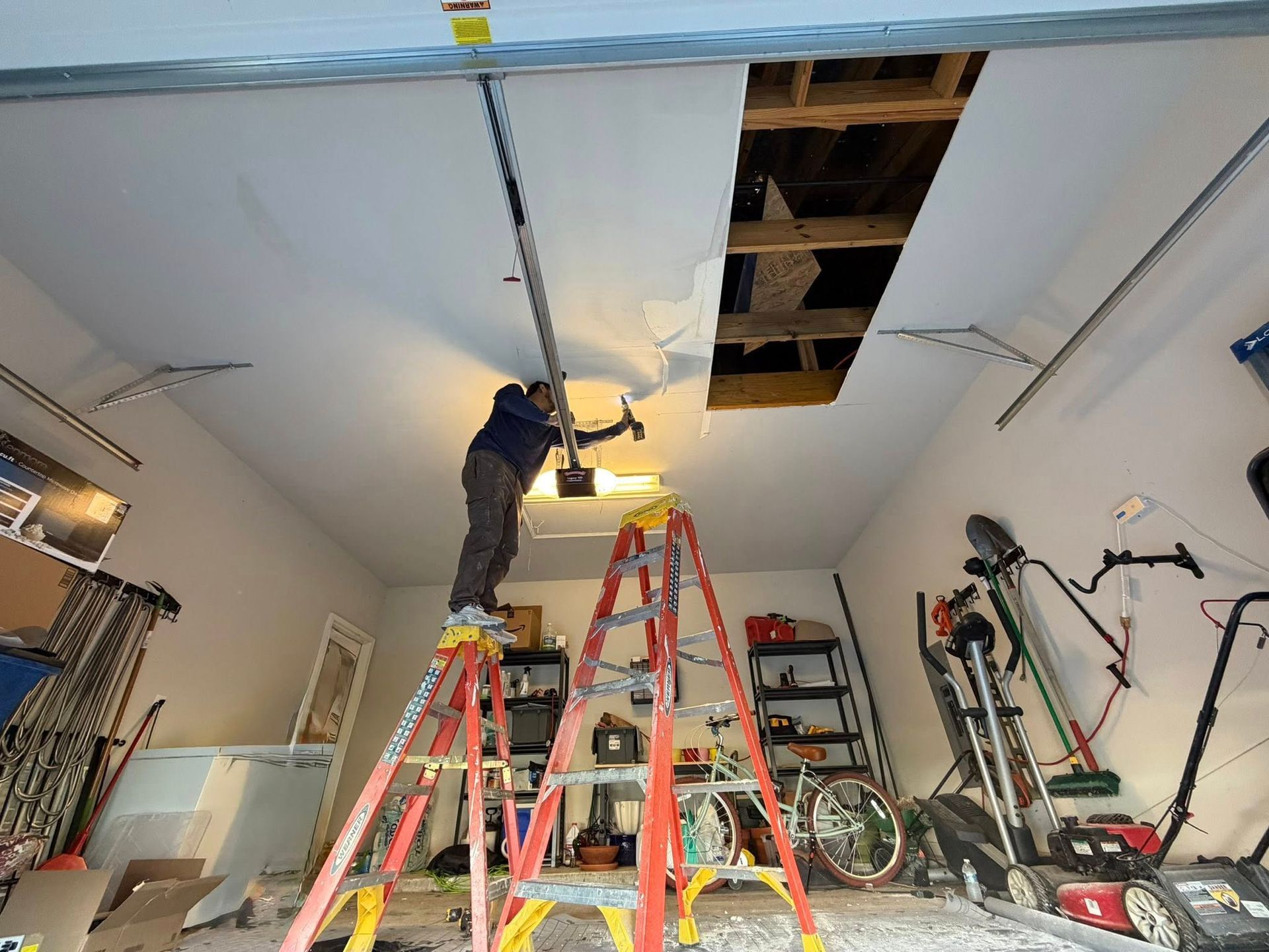 Worker standing on red ladder repairing ceiling in a cluttered garage with an open attic access panel.