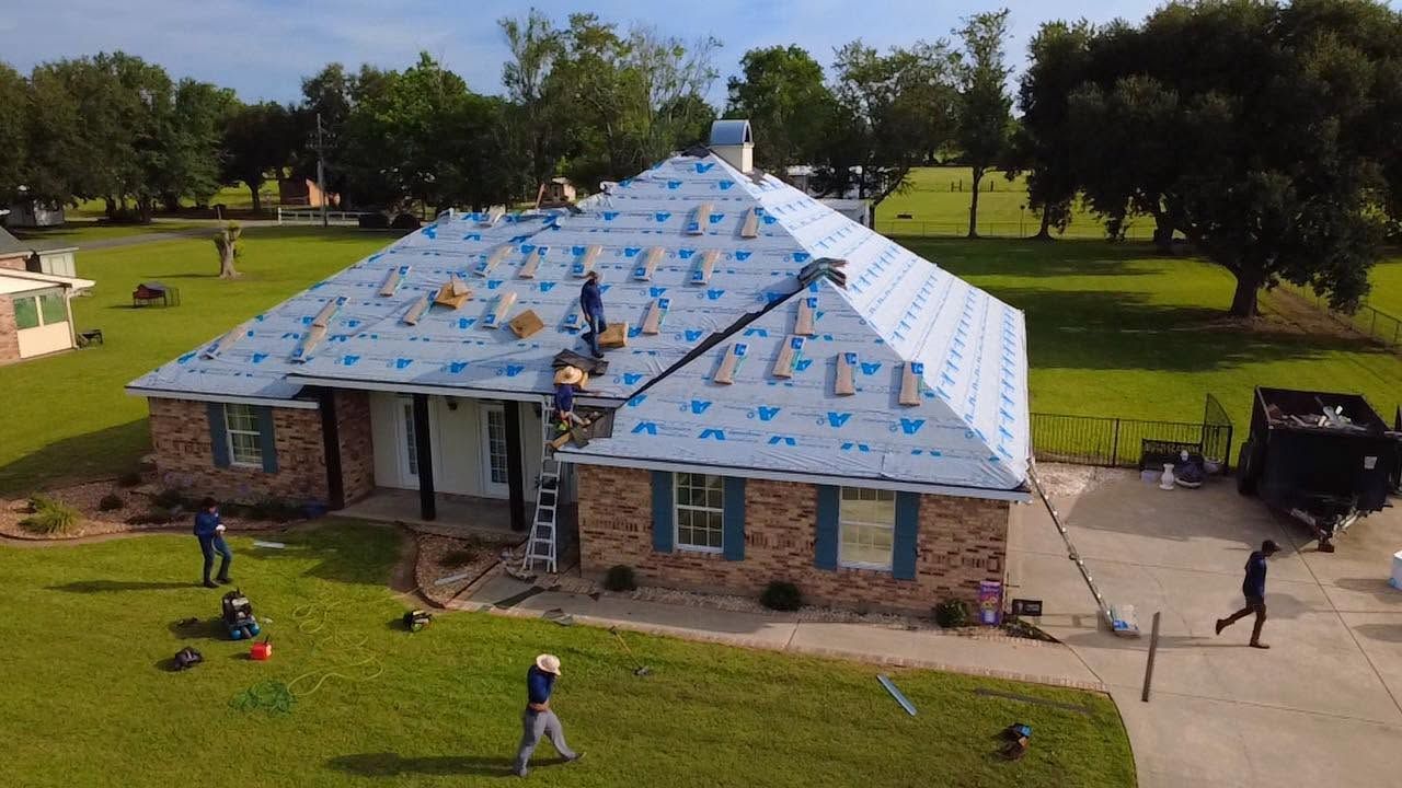 House with blue tarp roof in a grassy yard, with a few people nearby and trees in the background