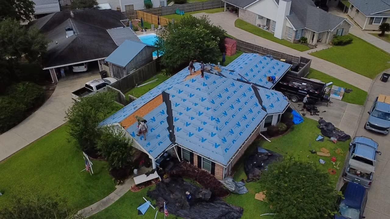 Aerial view of a house with a blue tarp-covered roof, surrounded by trees and neighboring homes.