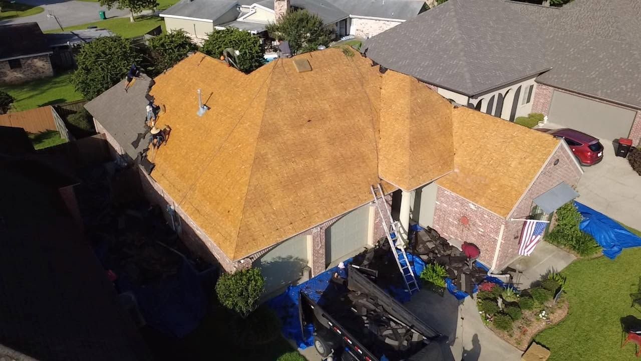 Aerial view of a house with a tan roof in a suburban neighborhood, with a car parked in the driveway.