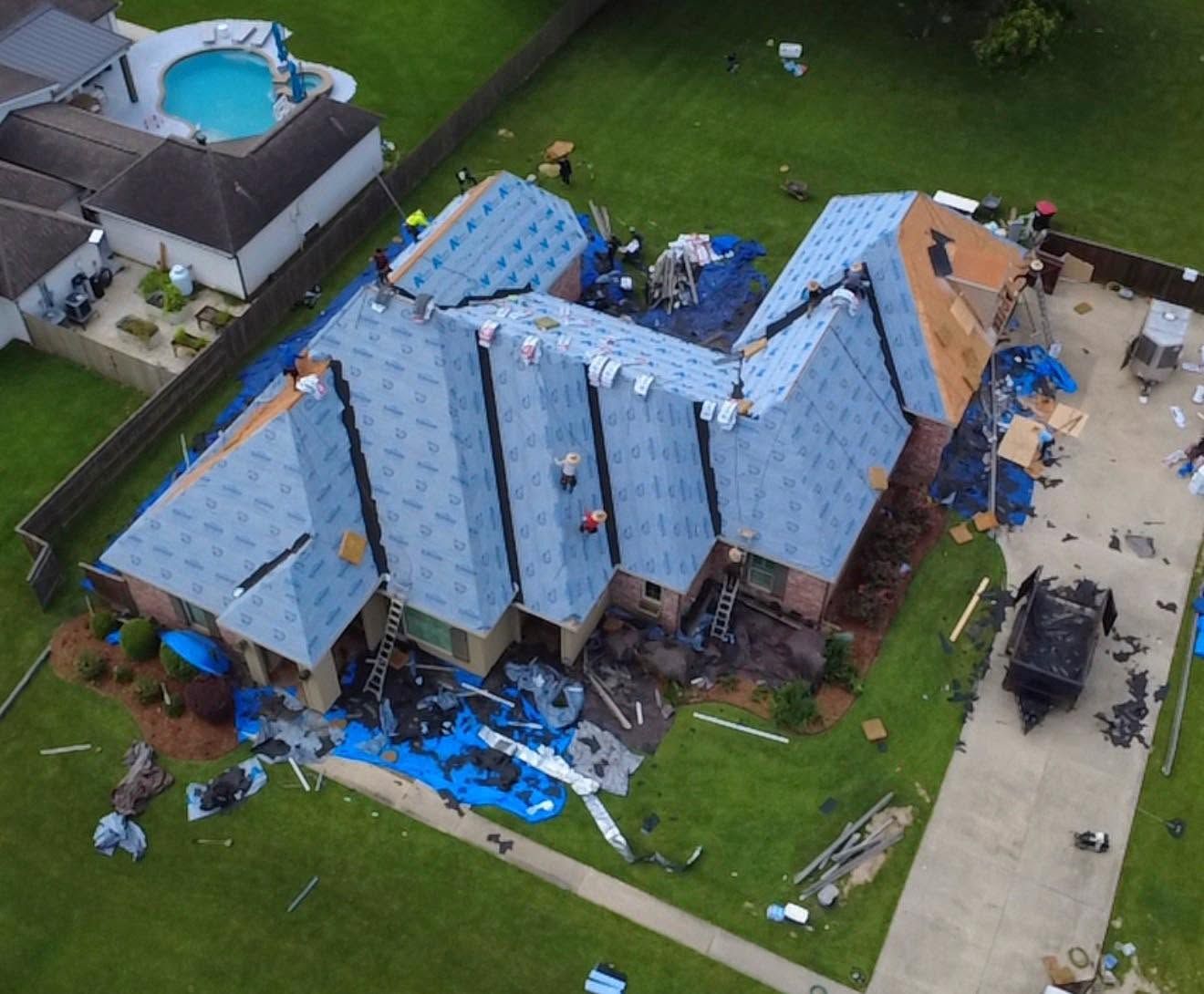 Aerial view of a house with a blue tarp roof after storm damage, with debris scattered around the yard.