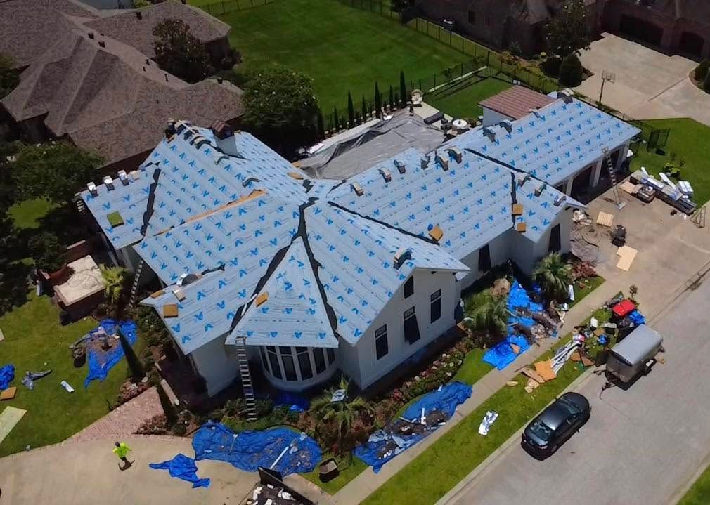 A house with a blue tarp-covered roof amid storm damage, with debris and vehicles nearby.