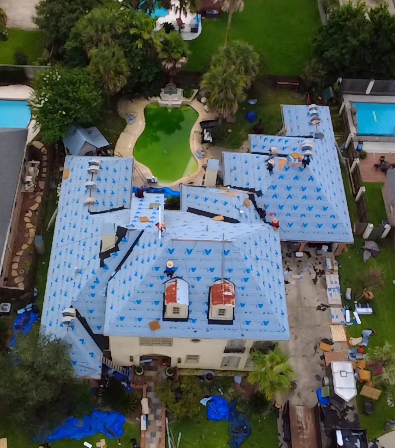 Aerial view of a house with a blue roof under repair, surrounded by trees and neighboring homes.