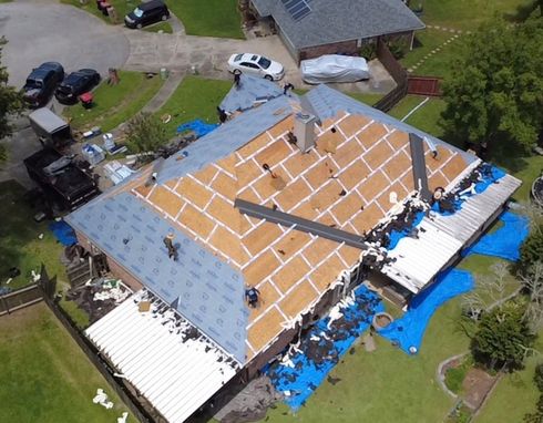 Aerial view of a house roof under repair, covered with plywood, tarps, and scattered construction materials.