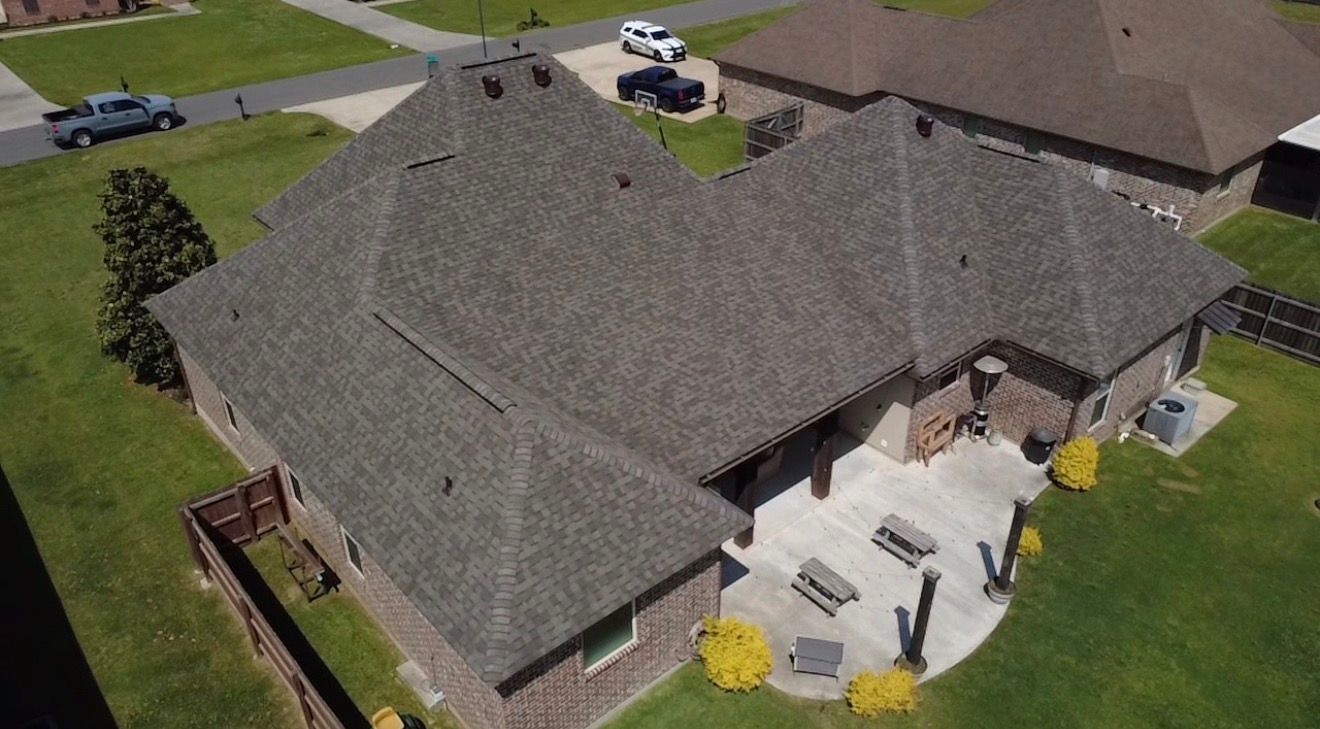 Aerial view of a suburban house with a gray shingle roof and backyard patio in a green lawn.