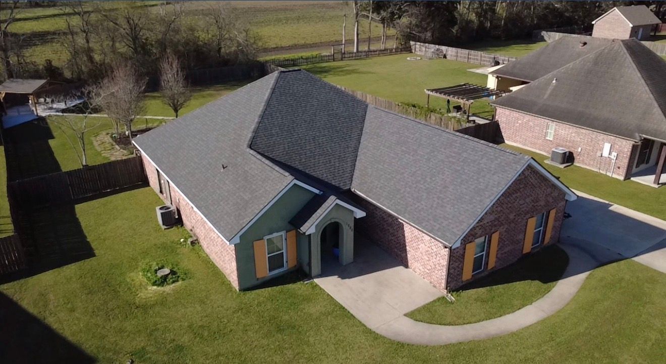 Aerial view of a brick house with a gray roof, green lawn, and curved driveway in a suburban neighborhood