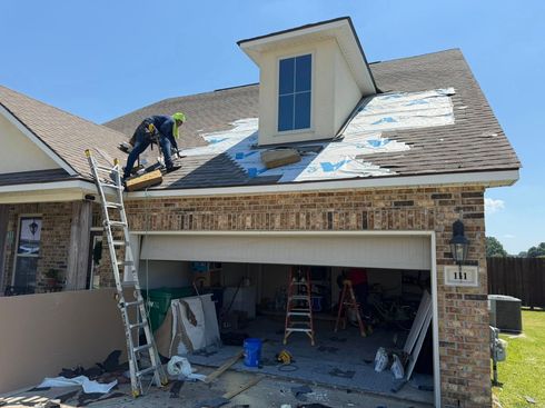 Worker on a roof above an open garage, with ladders and roofing materials.