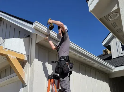 Worker on a ladder fastening trim to a house exterior under a clear blue sky