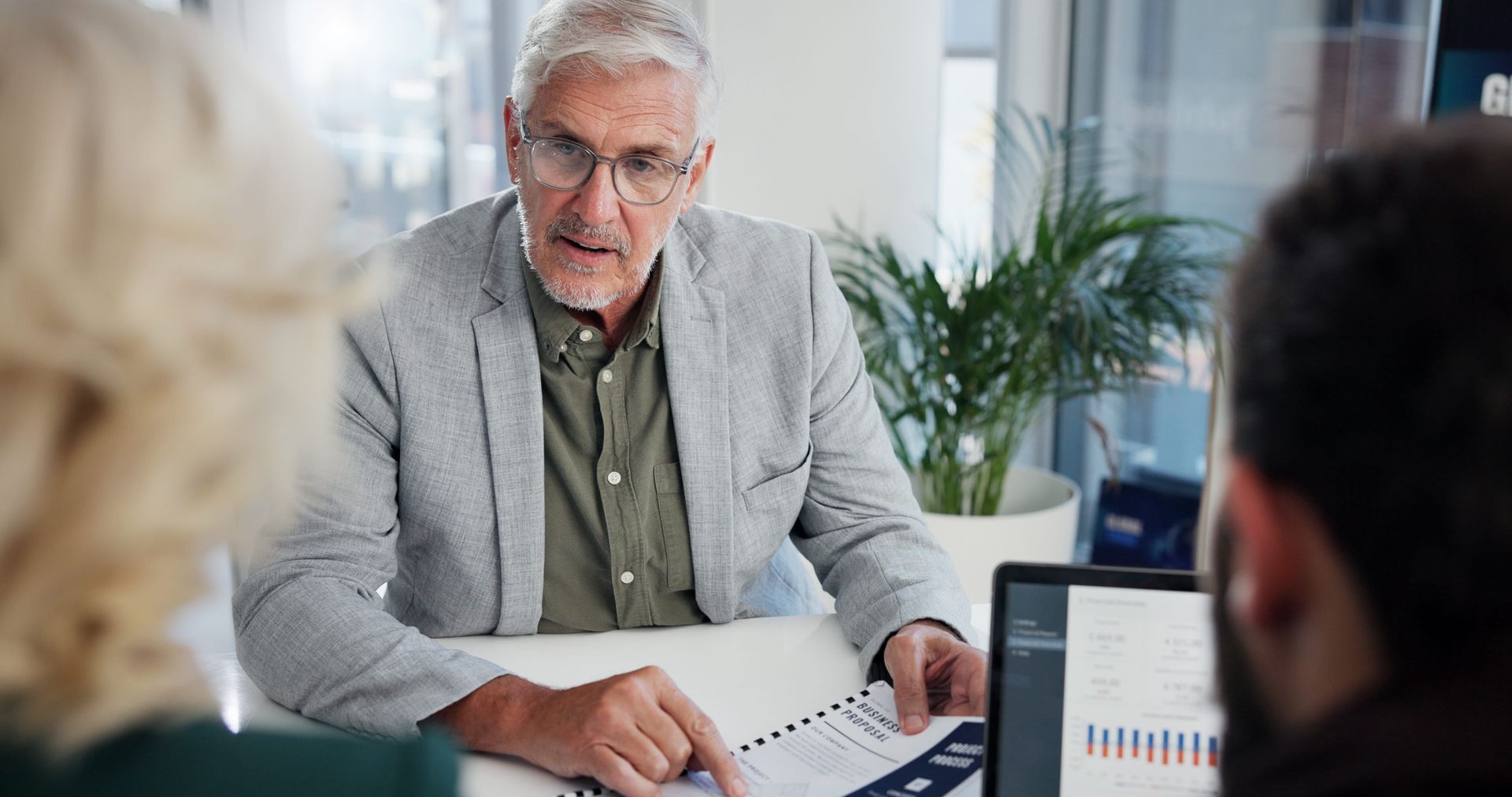 Older man in a gray blazer reviewing documents during a meeting with two people in an office