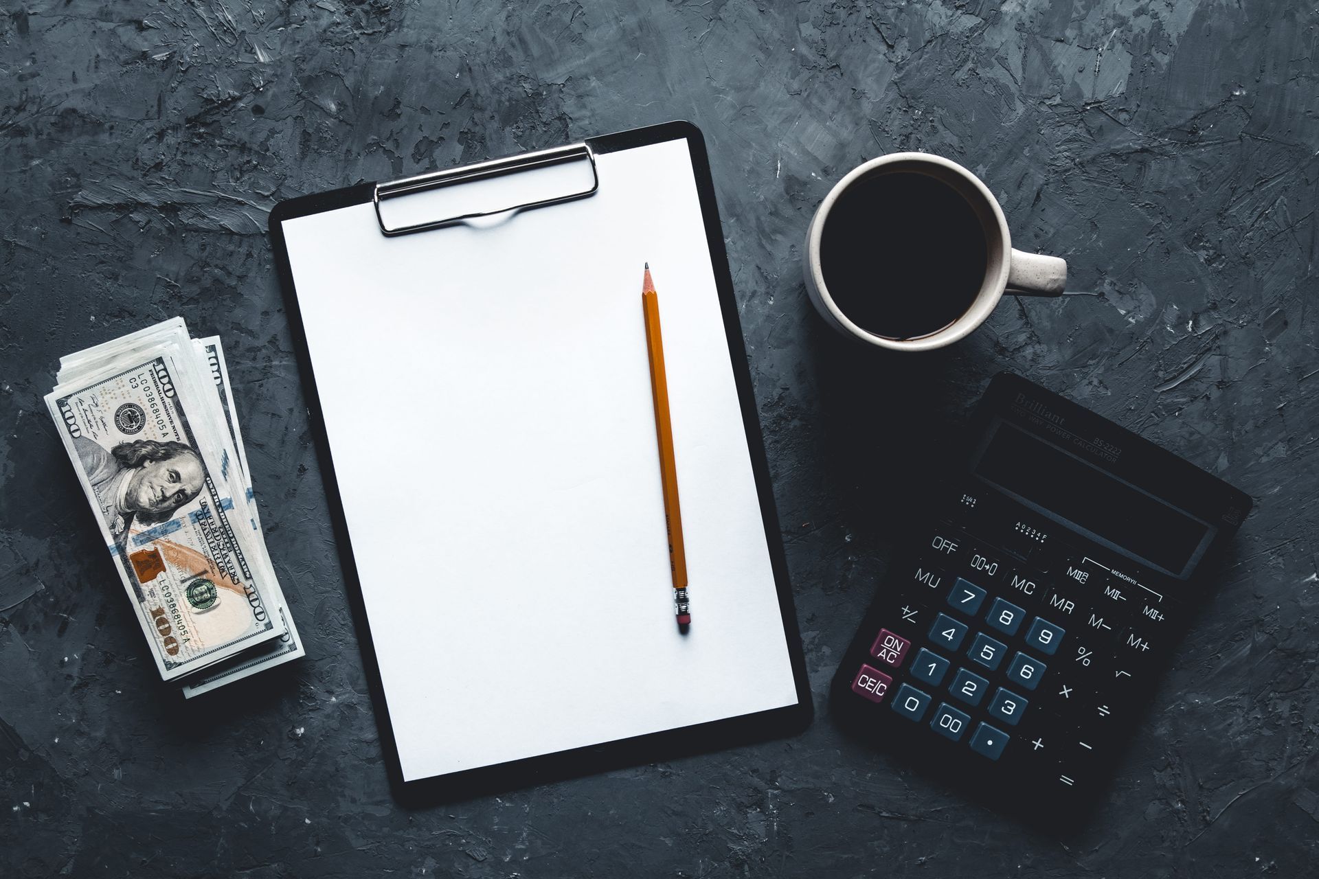 Clipboard with pencil, calculator, coffee cup, and cash on a dark desk.