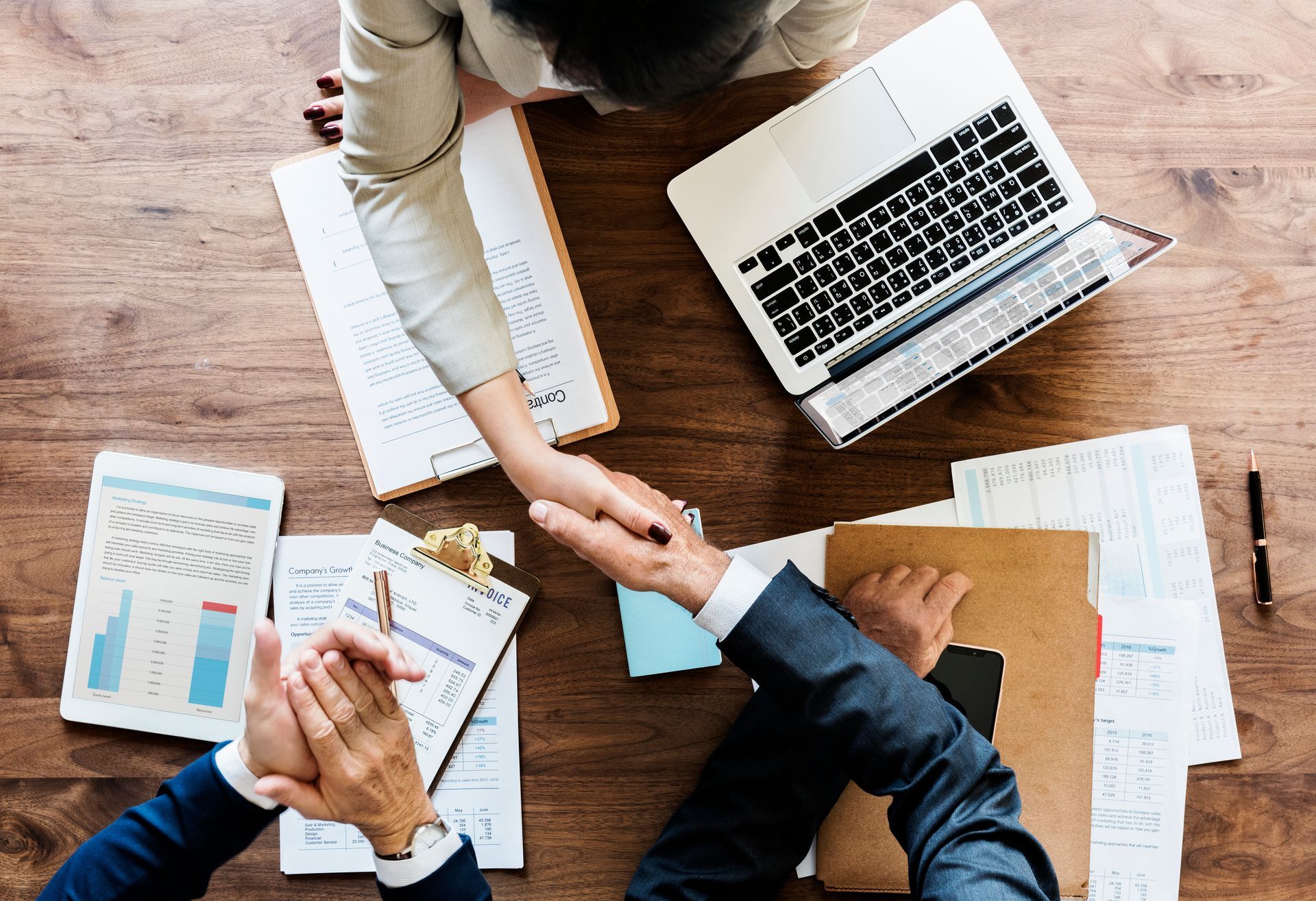 Three people in a meeting table, shaking hands over documents and a laptop.