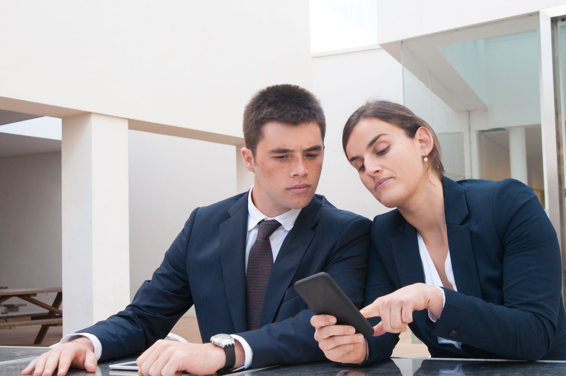 Two coworkers in suits reviewing a smartphone at a desk in a modern office