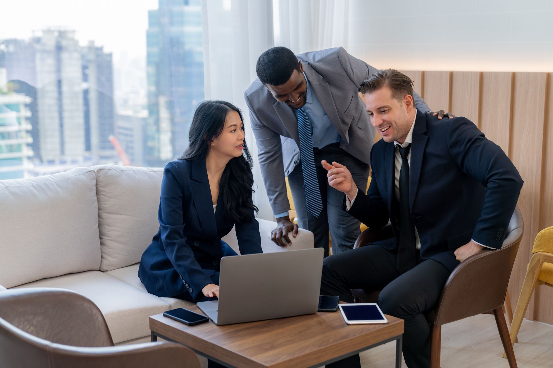 Three coworkers discussing a laptop in a modern office lounge with a city skyline behind them