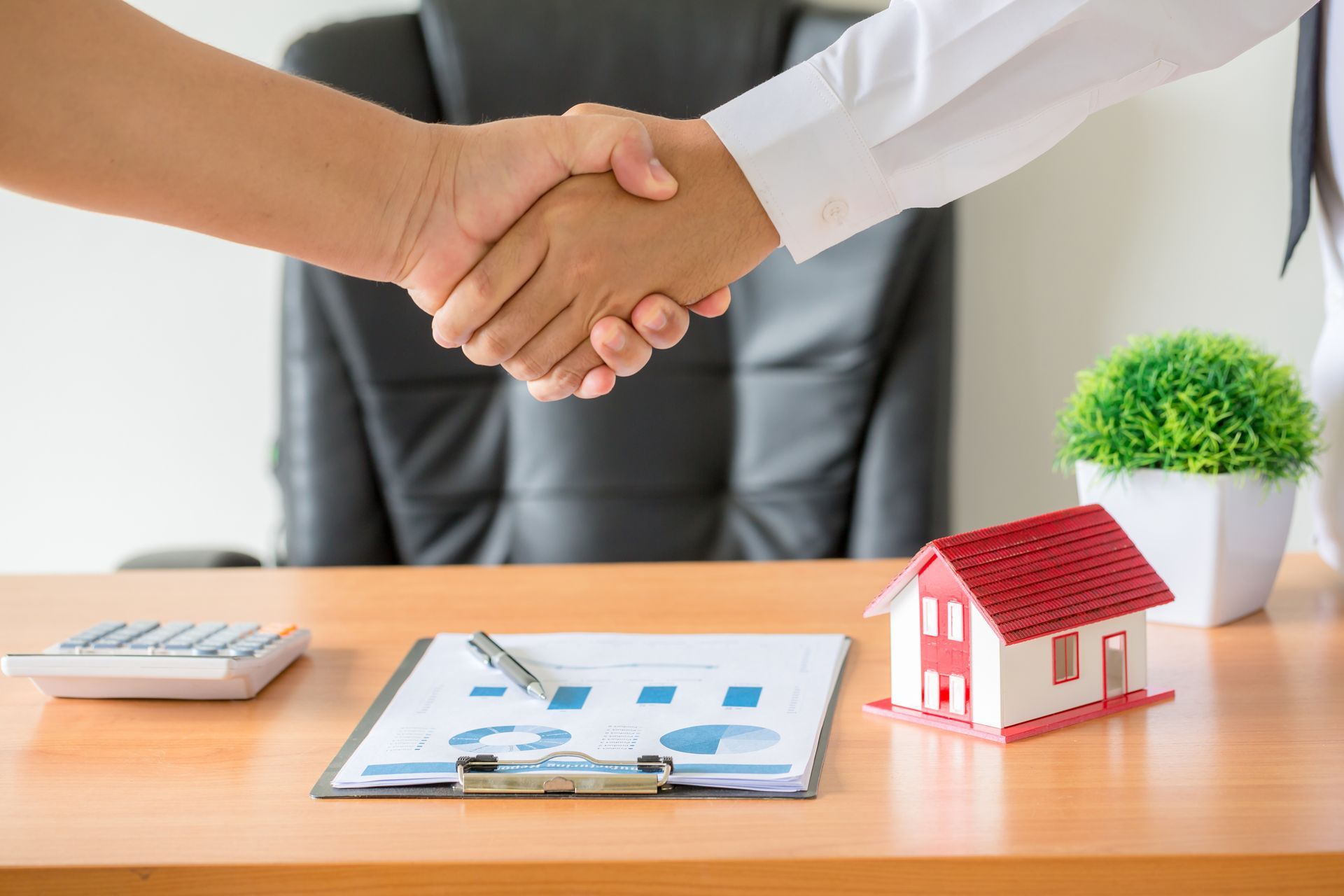 Handshake over office desk with house model, clipboard, calculator, and small plant
