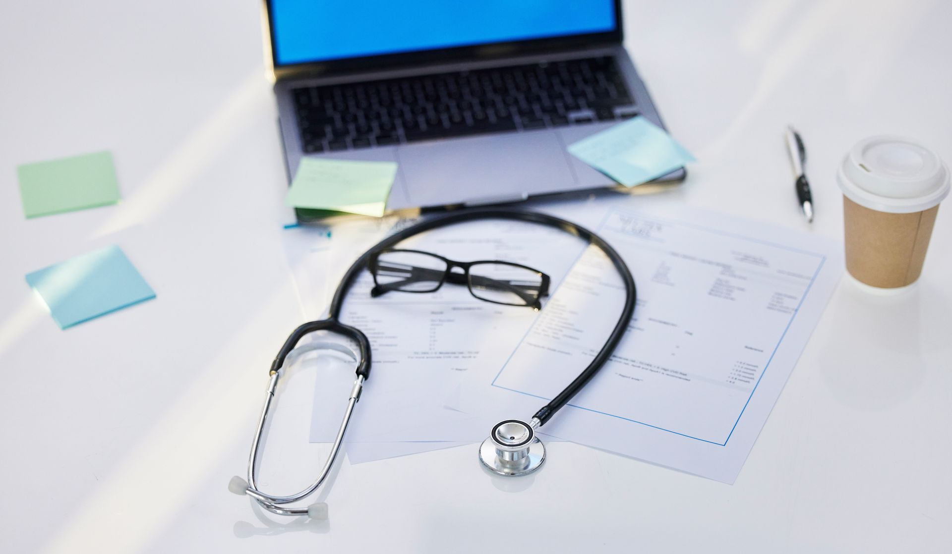 Medical stethoscope, eyeglasses, chart, coffee cup, and sticky notes on a desk with an open laptop.