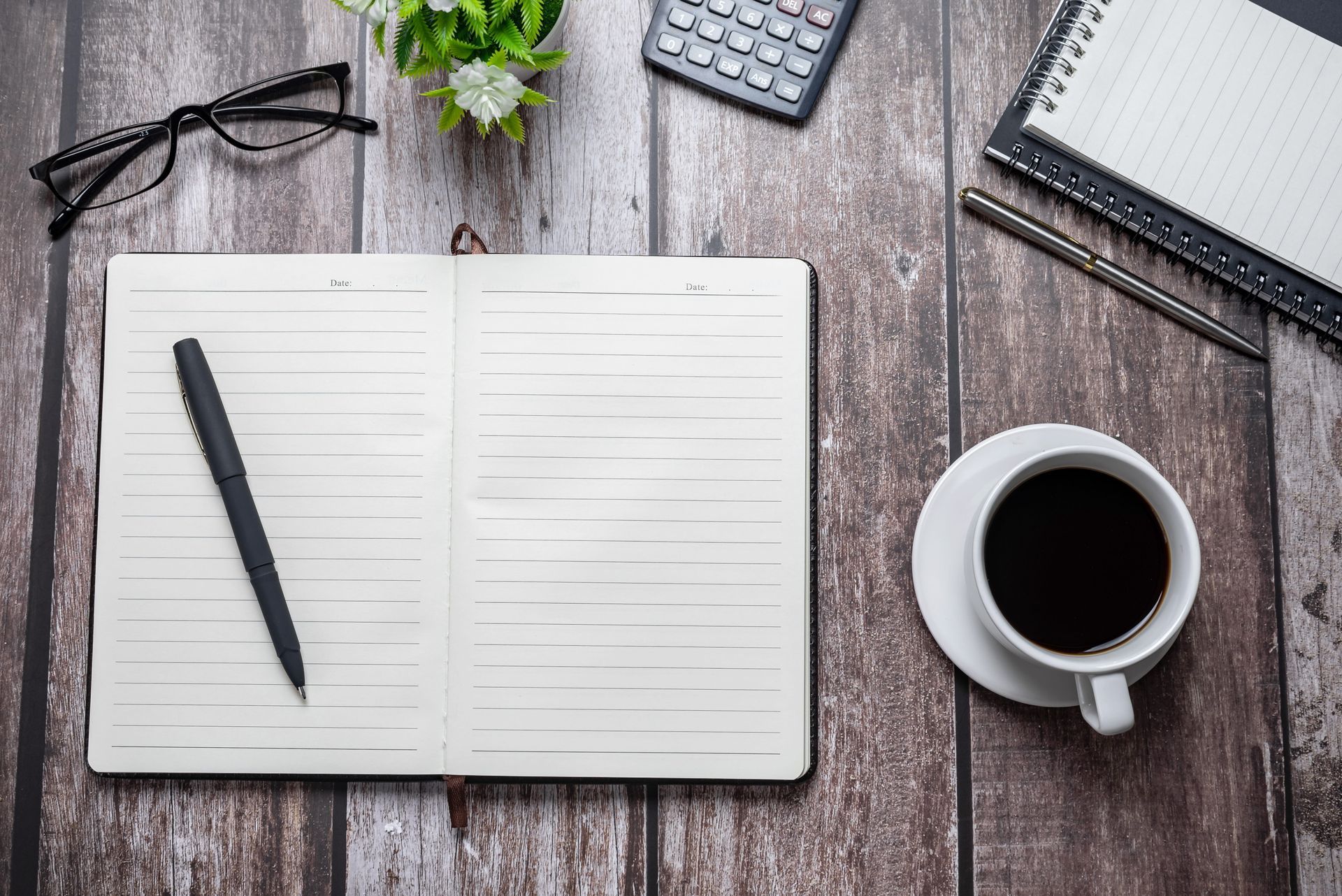 Open notebook with pen, coffee cup, glasses, calculator, and plant on a rustic wooden desk