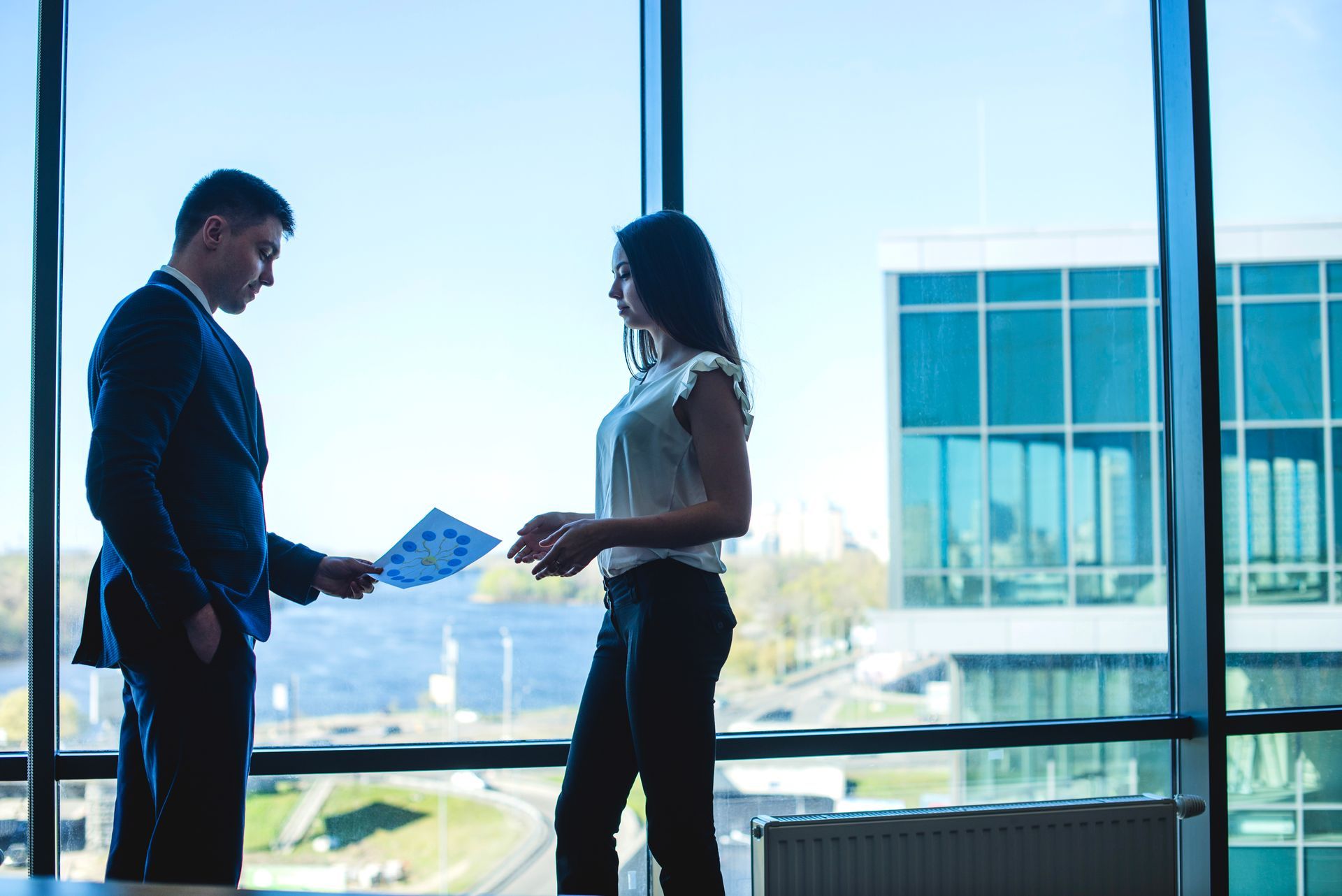 Two people talking by a large office window, holding papers, with a city view outside.