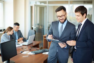 Two businessmen in suits reviewing a tablet in a bright office with coworkers and laptops in the background