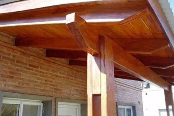 Techo de porche de madera con vigas a la vista sobre una pared de ladrillo y ventanas.
