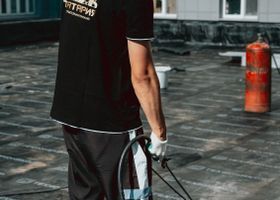 Una persona camina con su bicicleta por una calle mojada, vistiendo una camisa negra y guantes blancos.