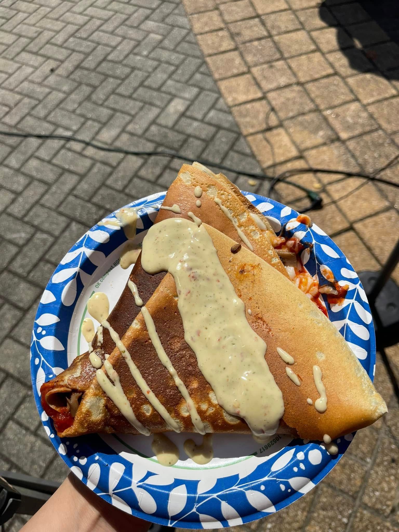 Paper plate with a toasted sandwich drizzled in sauce, held outdoors over a brick-paved walkway.