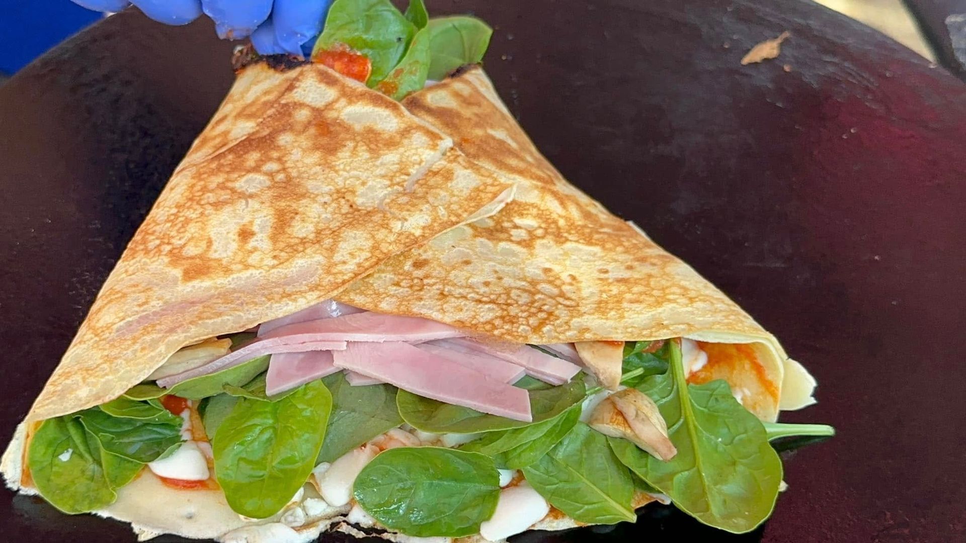 A folded crepe sandwich with greens and ham on a dark plate, held open by a gloved hand