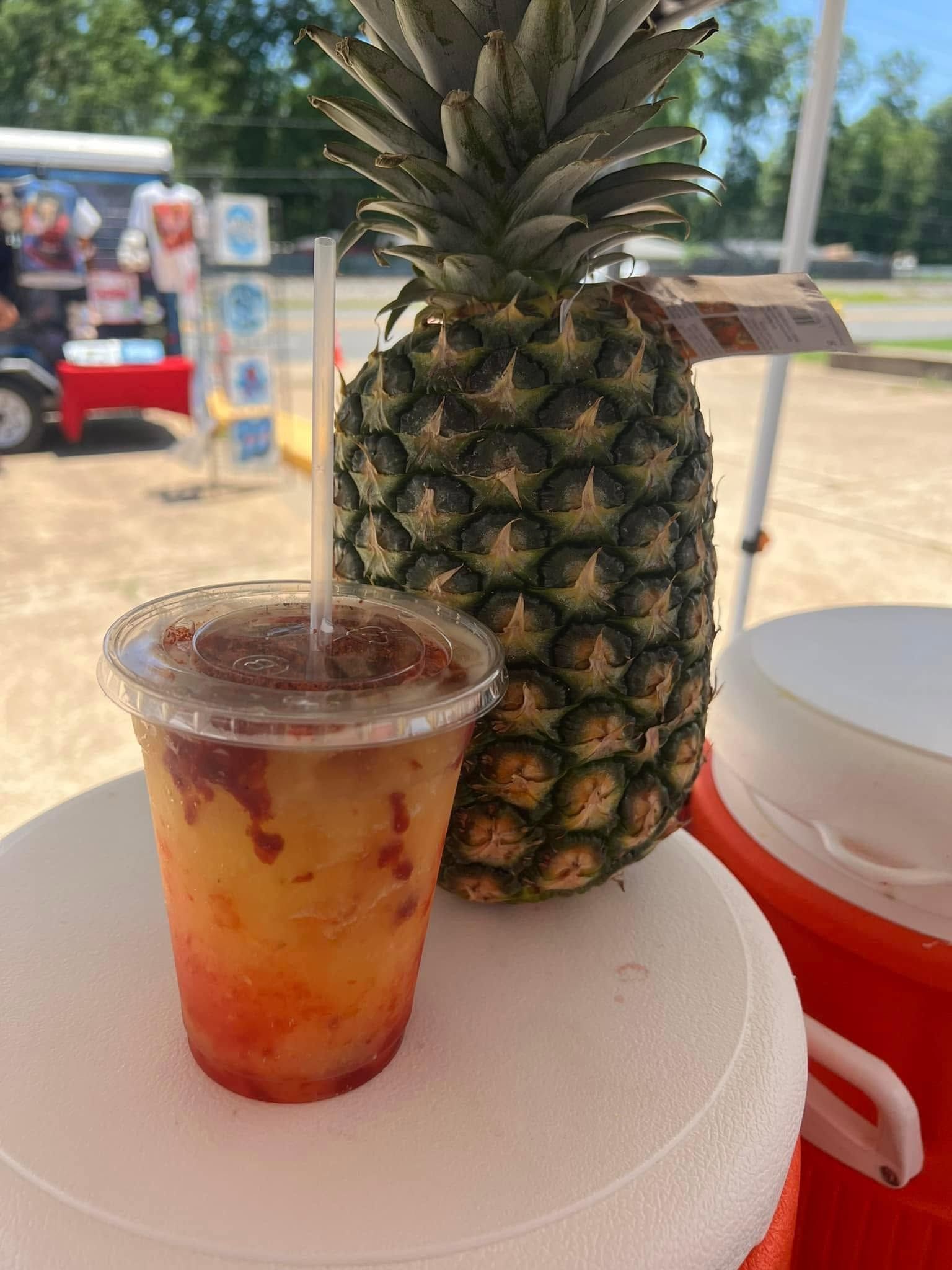 Pineapple drink in a clear cup beside a whole pineapple on a white table outdoors.
