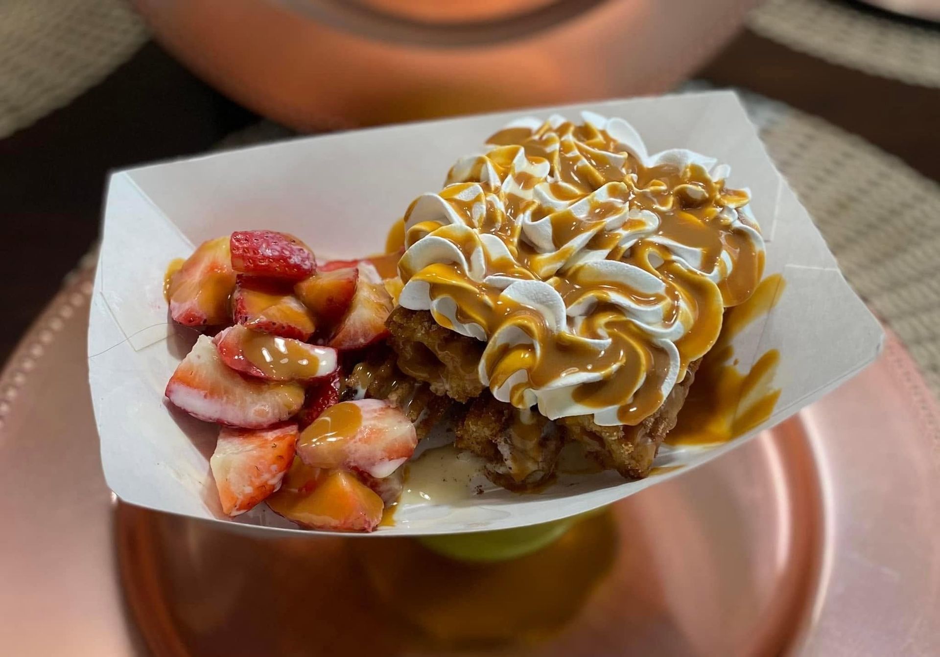 Waffle topped with whipped cream and caramel, served with sliced strawberries on a paper tray
