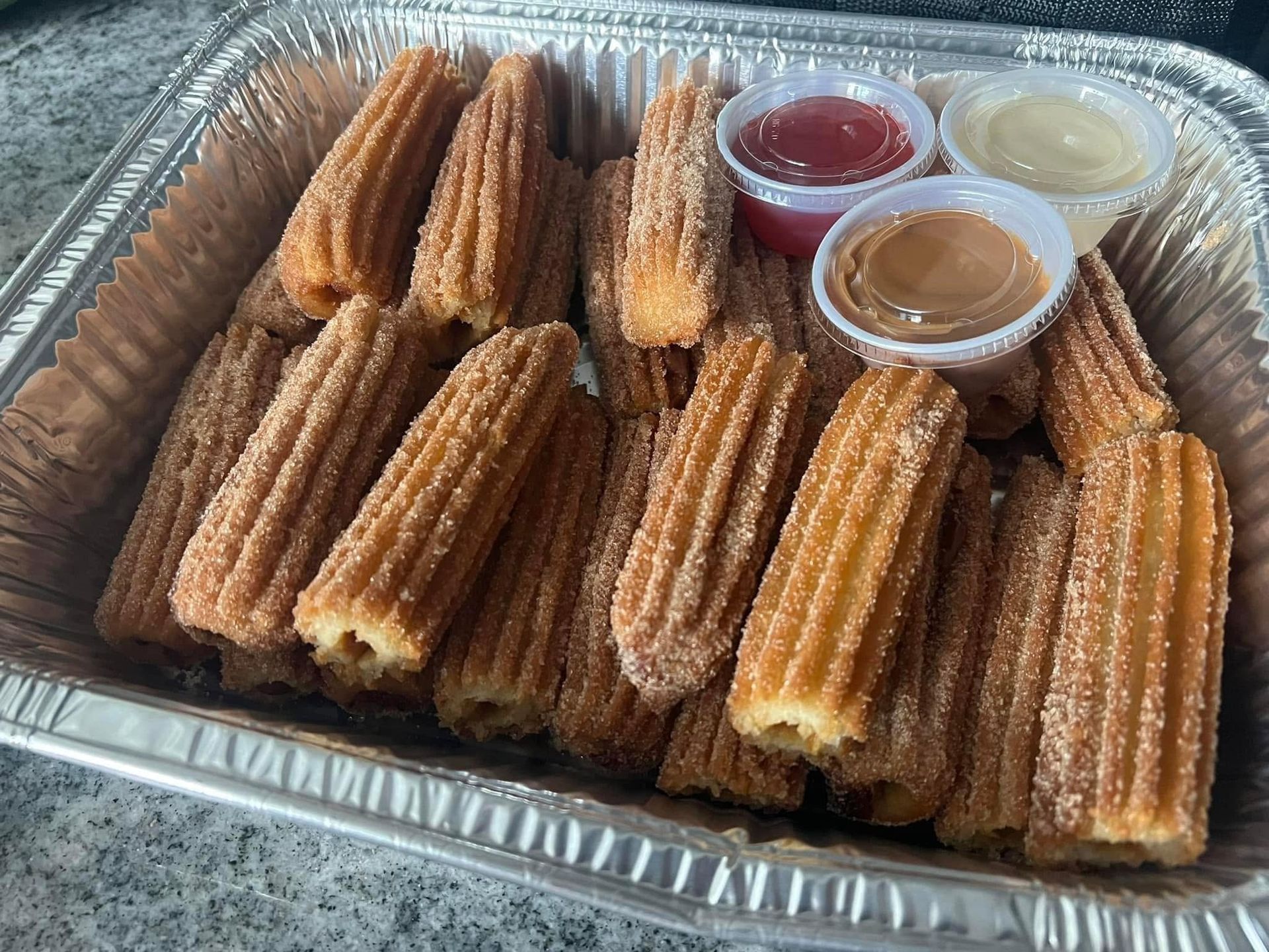 Tray of churros with three dipping sauces in a foil container
