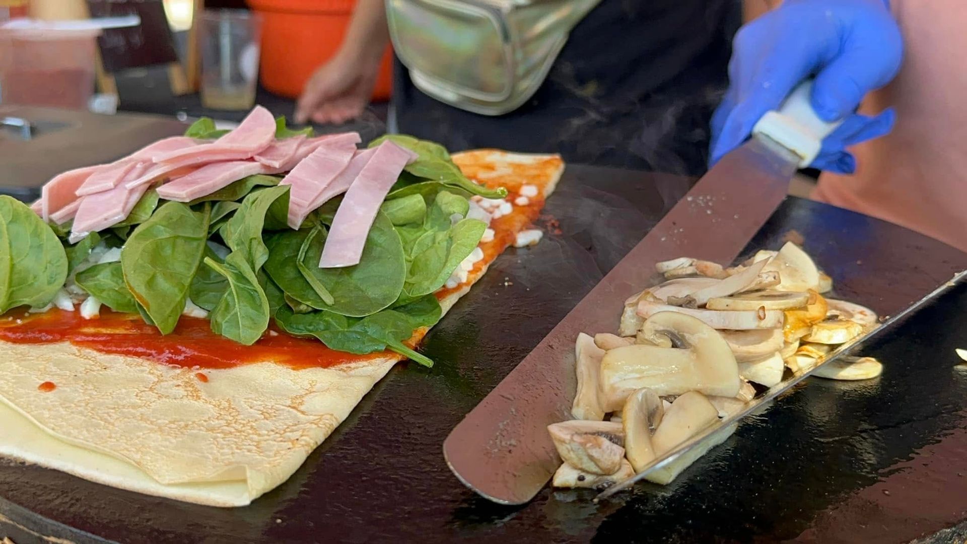 Cook slicing mushrooms on a black griddle beside an open sandwich with greens and meat