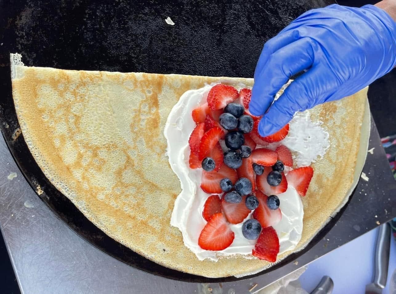 Hand placing blueberries on a crepe topped with whipped cream and sliced strawberries
