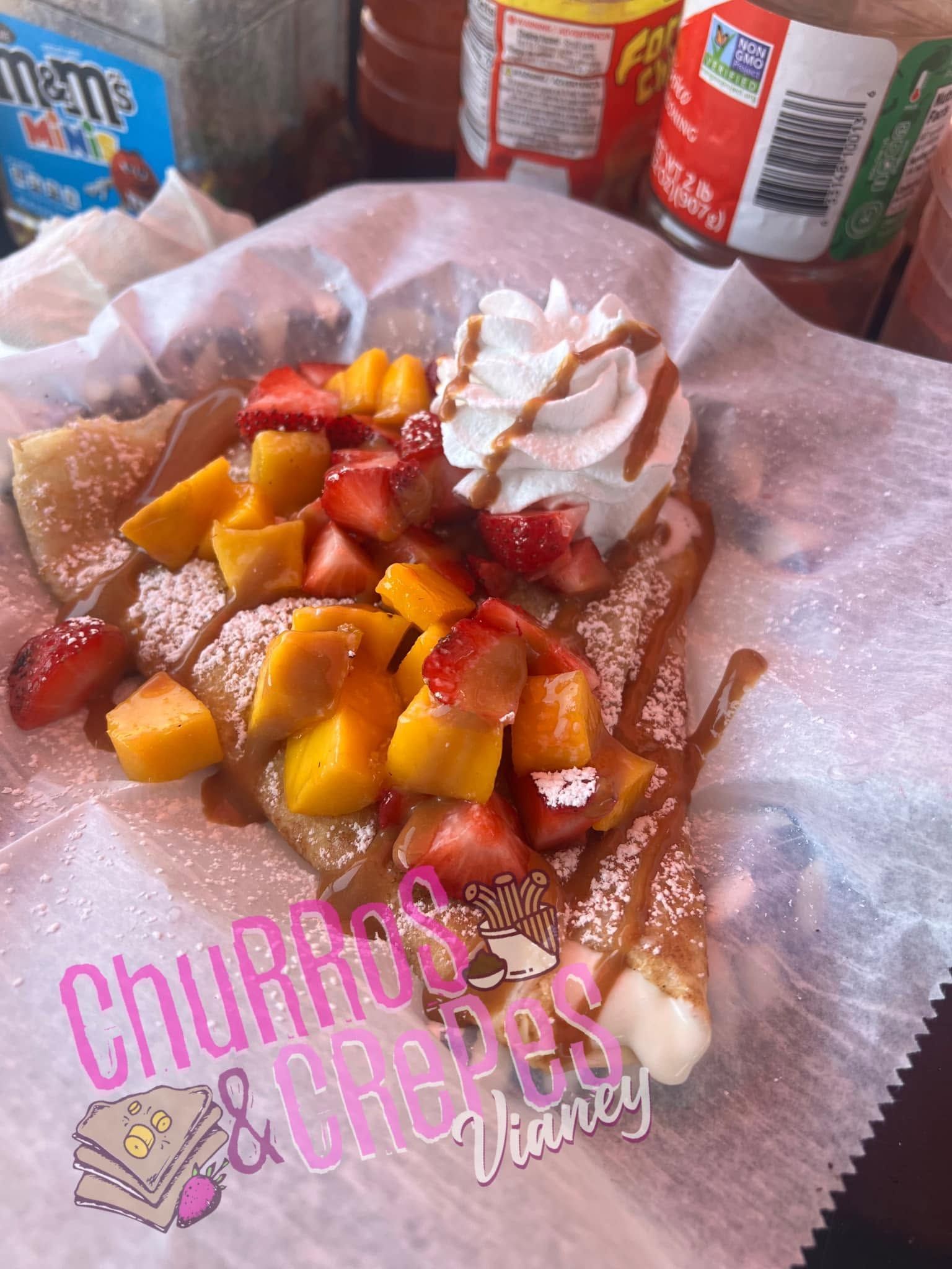 Churros topped with diced strawberries, peaches, and whipped cream on pink paper, with soda cans in the background