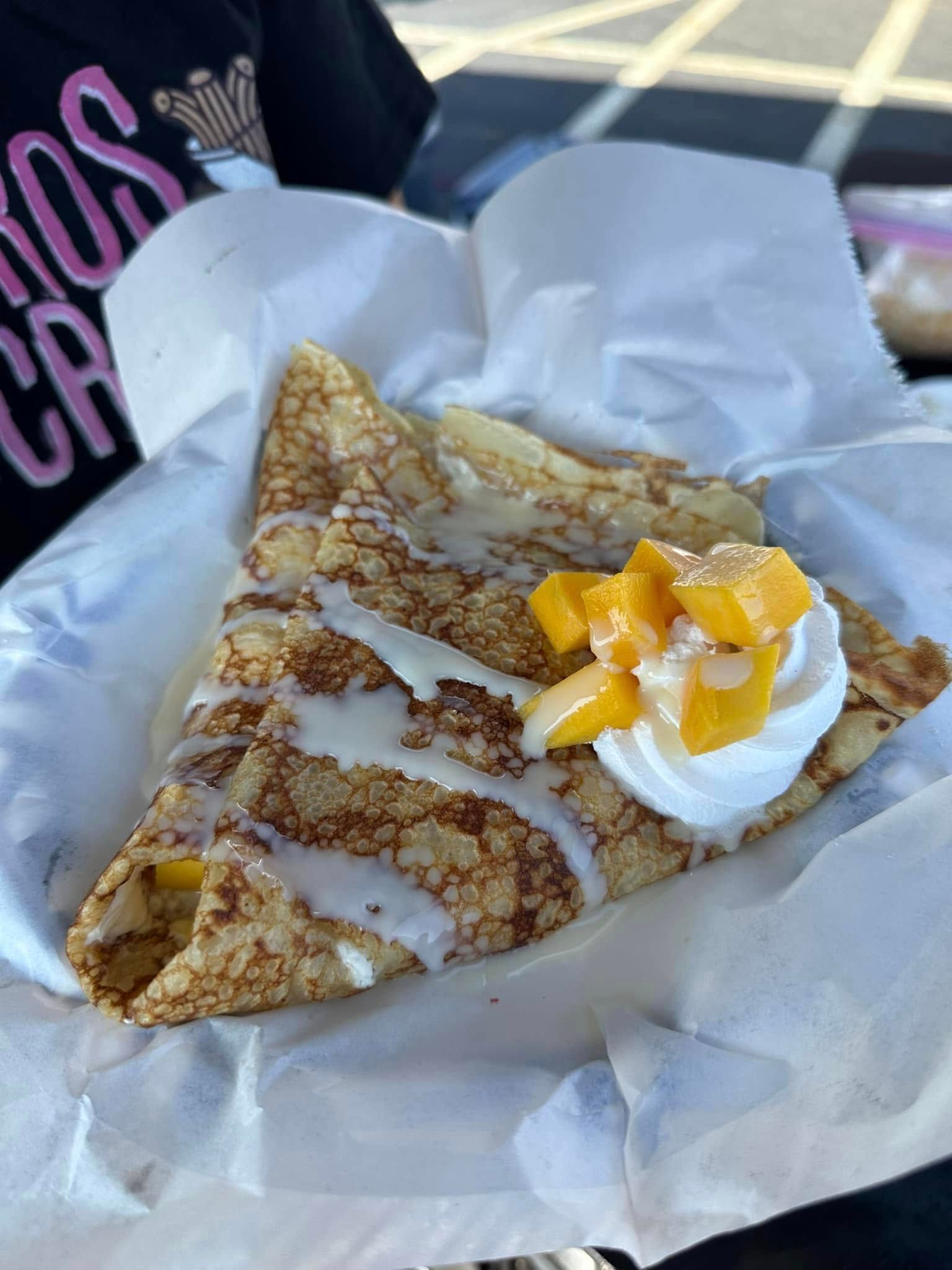 Crêpe topped with whipped cream and peach slices, held in white paper wrapping outdoors