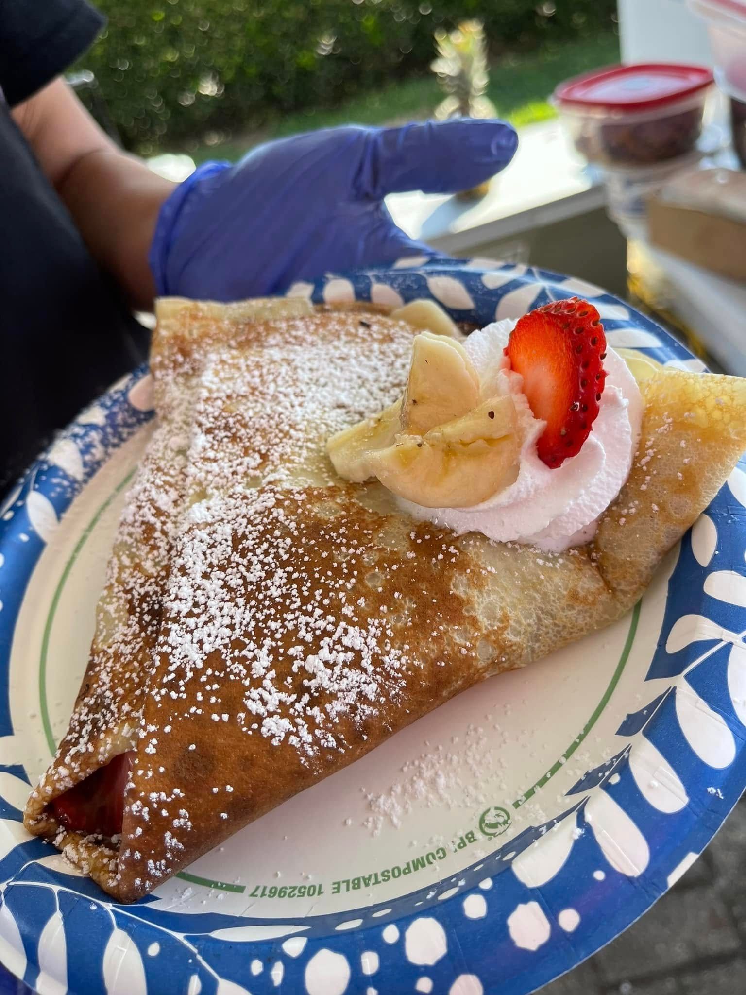 Crepe topped with whipped cream, strawberry slices, and powdered sugar on a paper plate