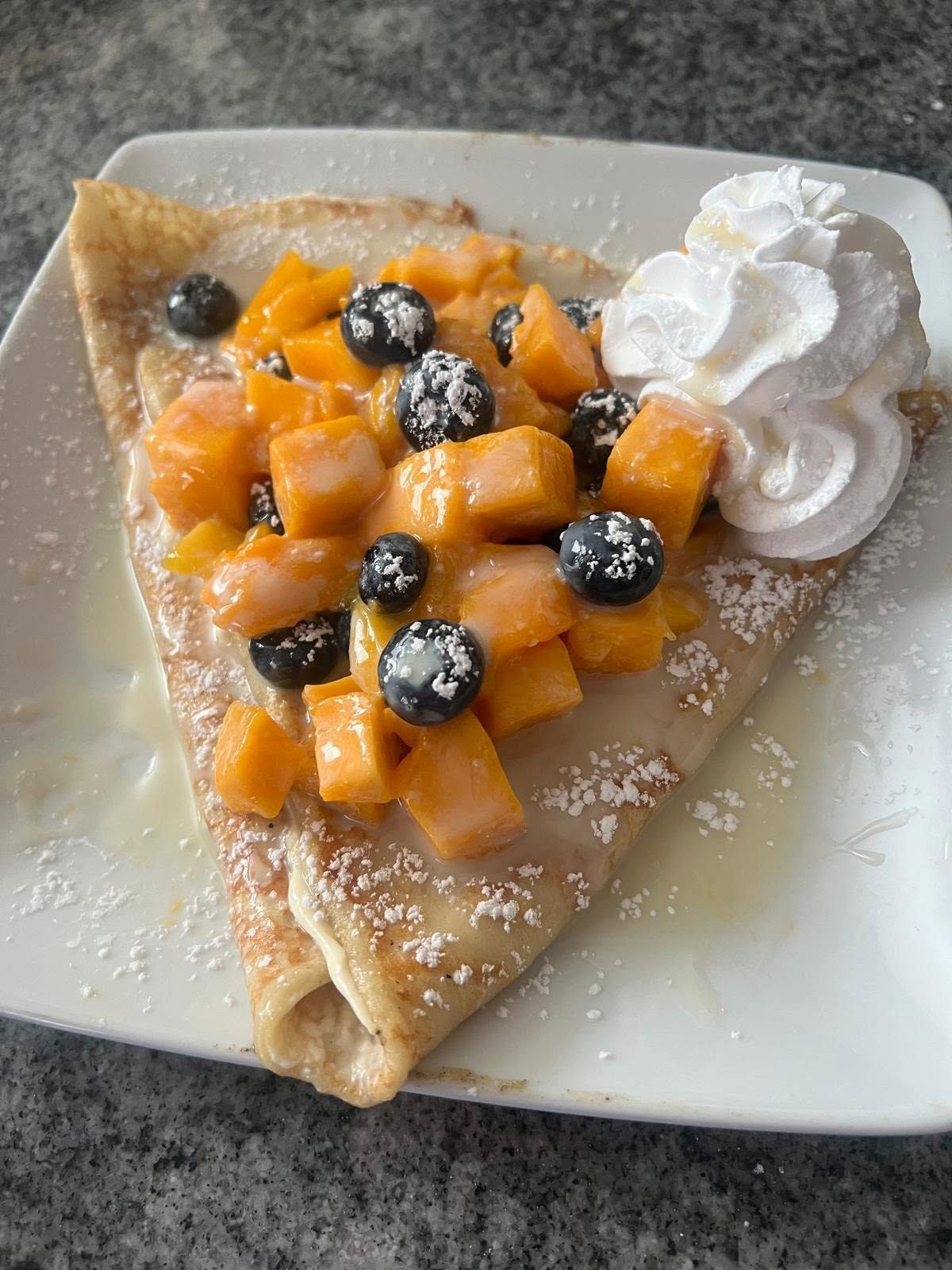 Crepe topped with diced peaches, blueberries, powdered sugar, and whipped cream on a white plate