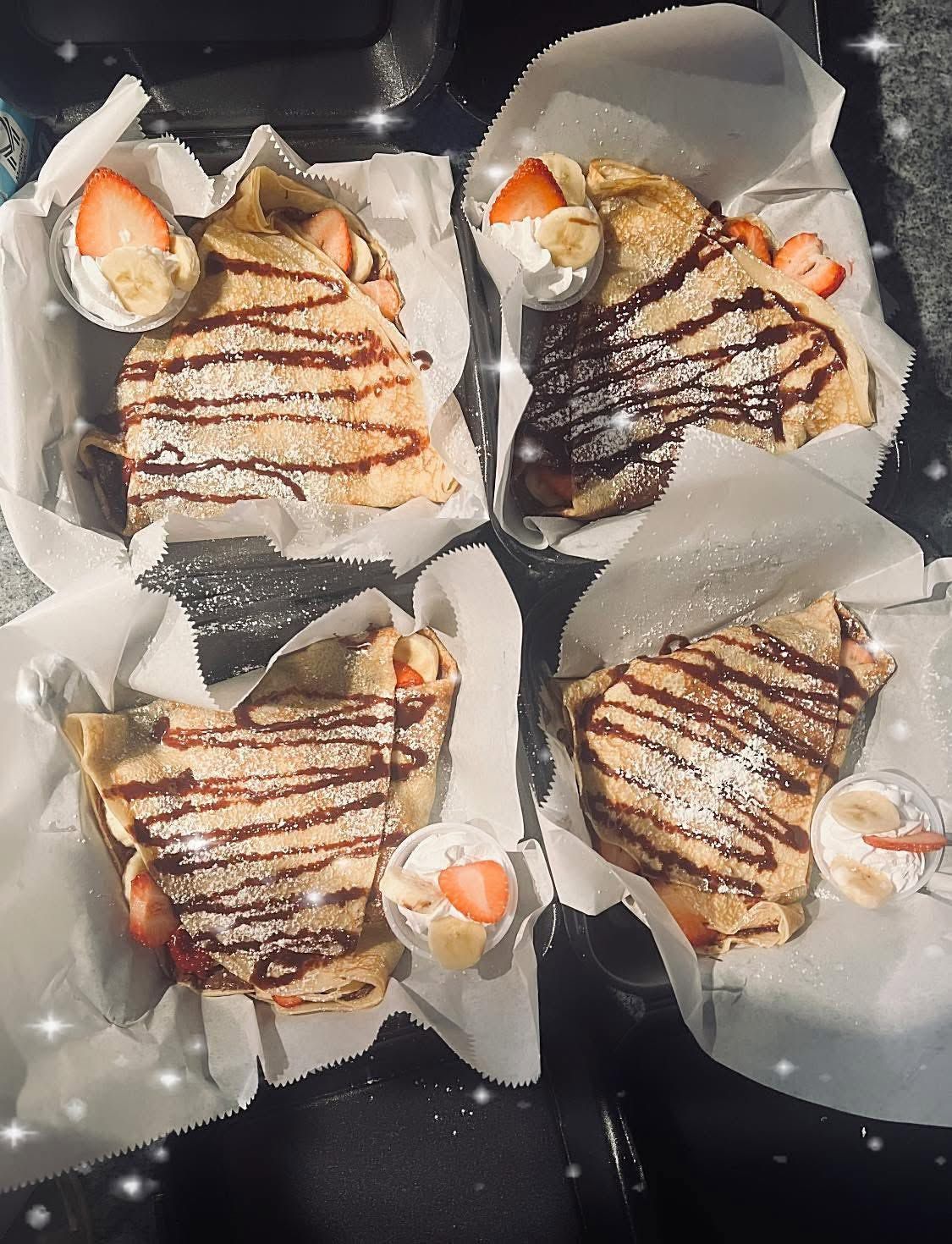 Four chocolate-drizzled crepes in paper wraps, topped with orange slices on a dark tray.