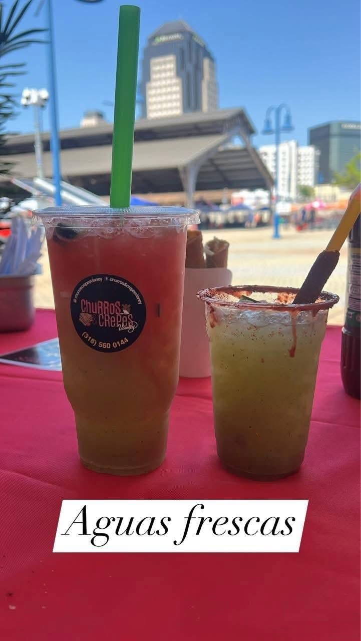 Two colorful aguas frescas in plastic cups on a pink table outdoors with a city skyline background