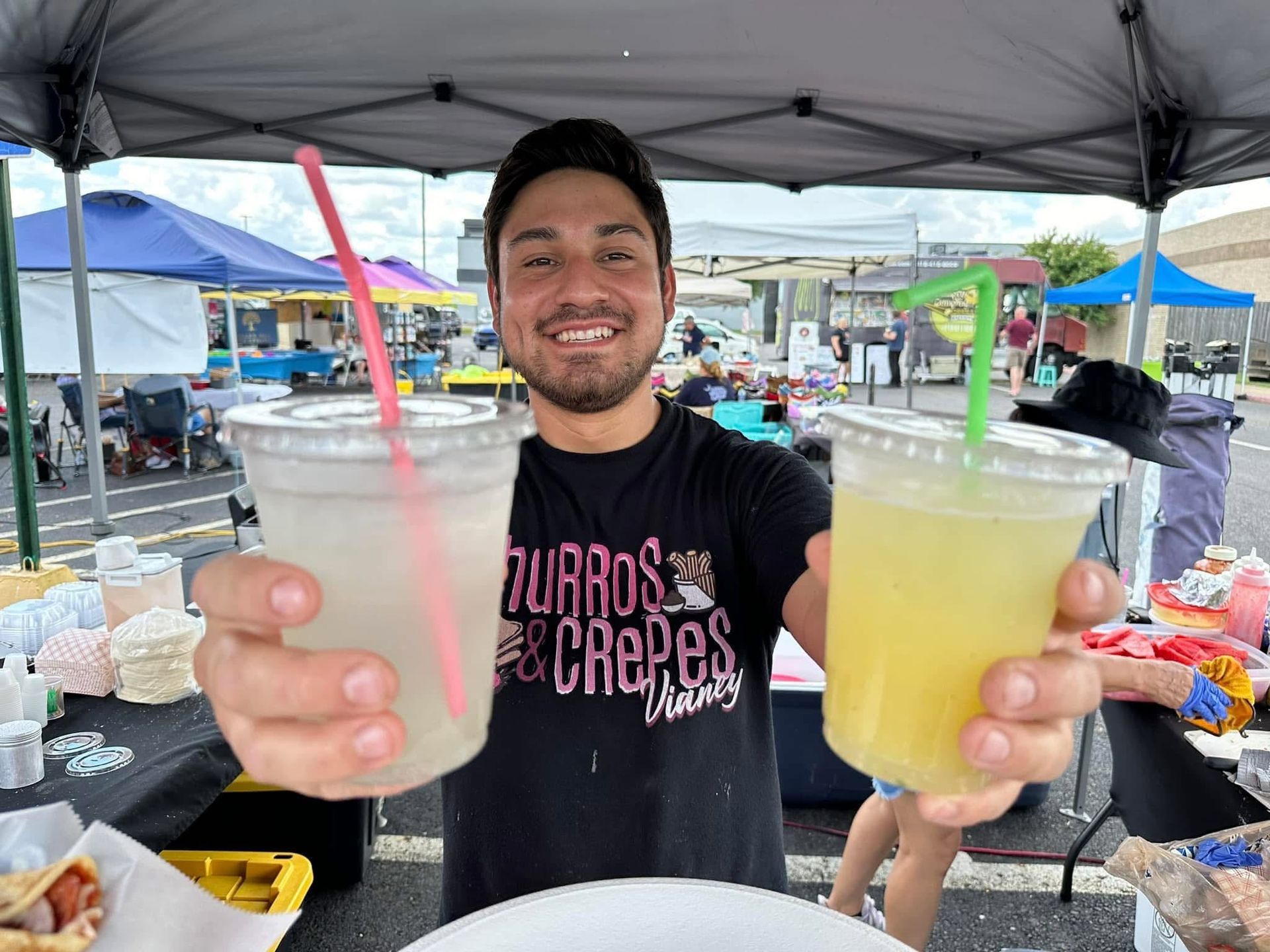 Smiling vendor holding two colorful drinks under a market canopy, with food stalls in the background