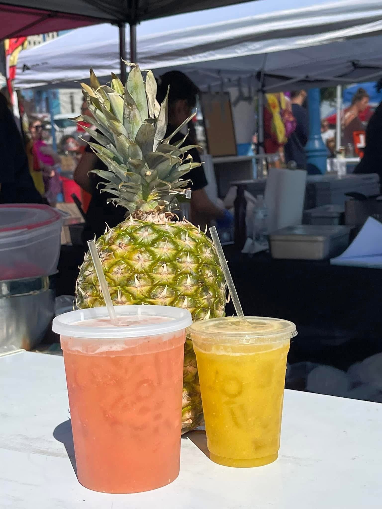 Two colorful drinks in plastic cups beside a pineapple at an outdoor market stall.