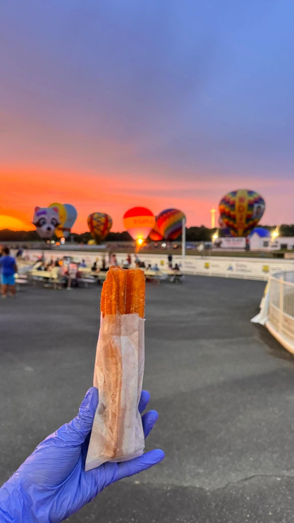 Blue-gloved hand holding a churro at sunset with colorful hot air balloons in the background